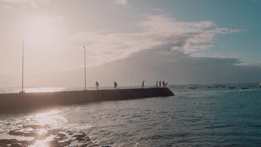 People walk and gather on a coastal pier during sunset with the ocean surrounding them