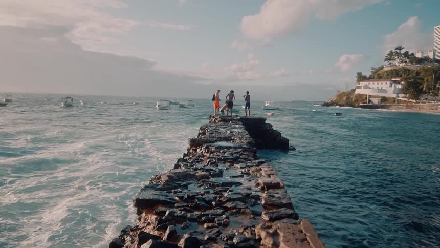Four people stand on a rocky path in the ocean as waves crash against the platform at midday