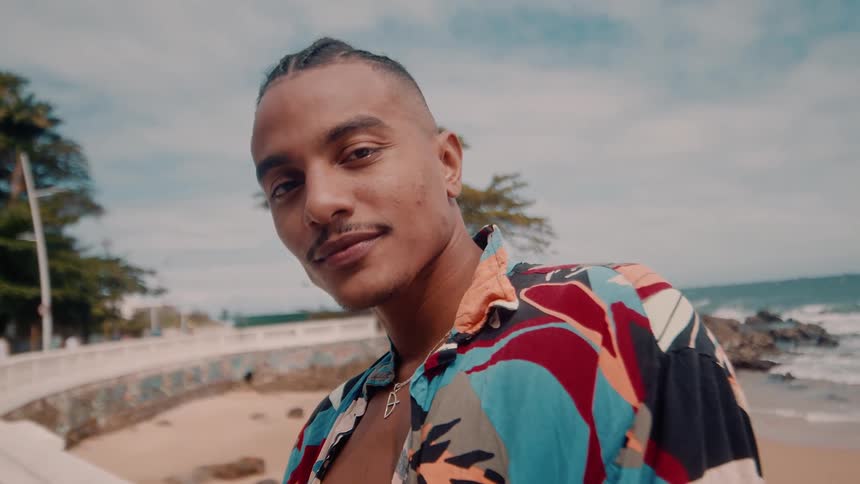 A smiling man stands near the beach in a colorful shirt looking directly at the camera