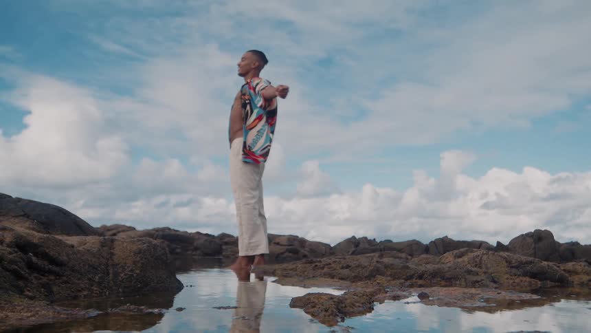 A man stands barefoot on coastal rocks with arms outstretched