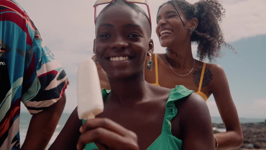 A smiling woman holding a popsicle at the beach shore with a man beside her and behind her another smiling woman also holding a popsicle