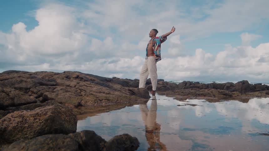 A man stands on rocky coastal terrain with arms raised under a bright sky