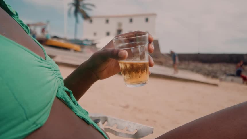A woman in a green swimsuit sits on a sandy beach holding a drink with palm trees and a white coastal building in the background