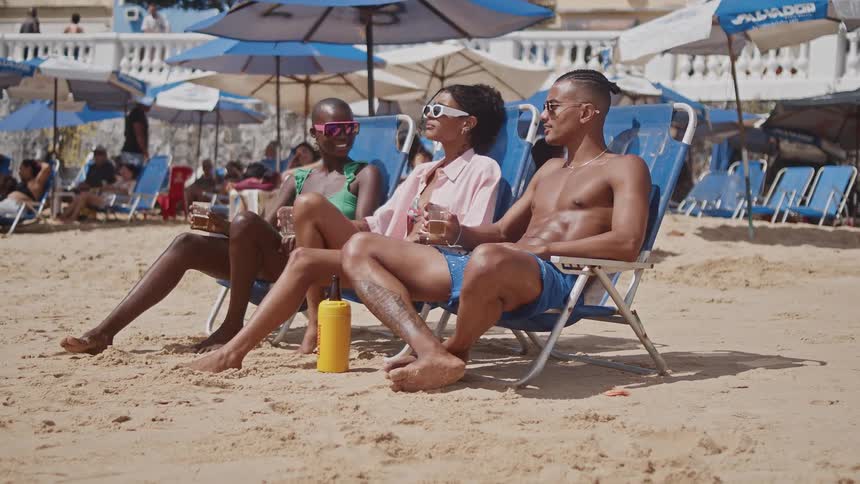 Three friends relax and enjoy drinks in lounge chairs on a bustling sun drenched beach surrounded by umbrellas