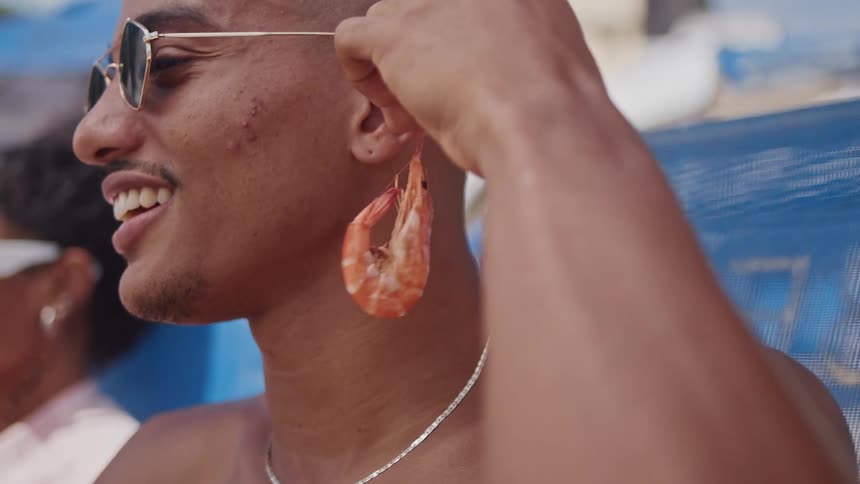 A young man in sunglasses playfully holds a shrimp to his ear like an earring at the beach
