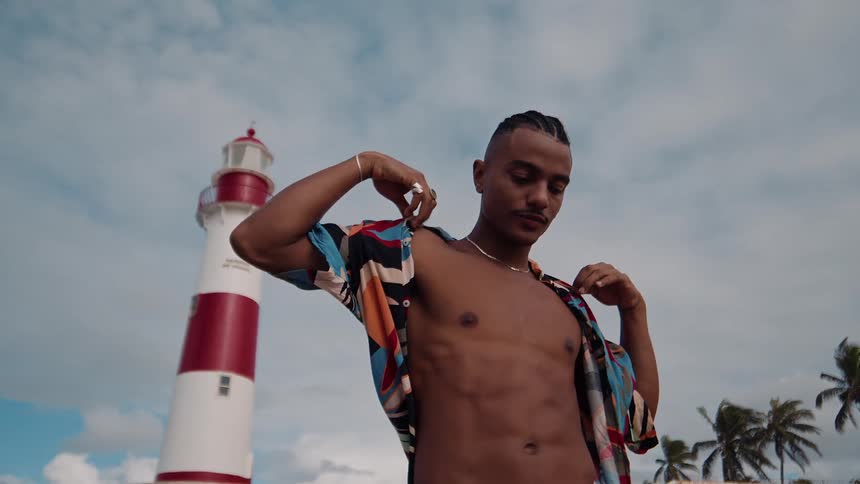 A young black man wearing an unbuttoned summer shirt stands beside a lighthouse