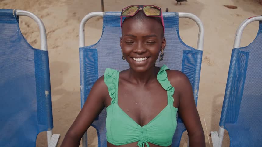 A smiling woman in a green bikini is sitting on a blue chair on the sand of a beach