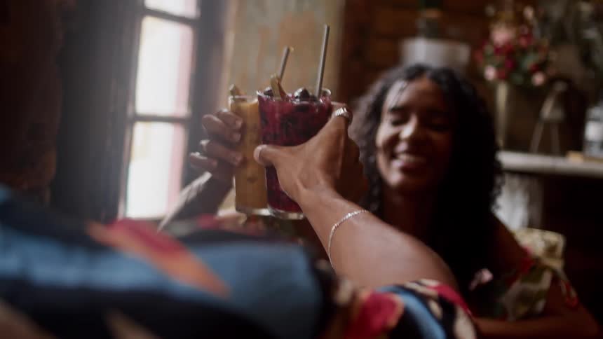 Two people joyfully clink colorful cocktails in a cozy indoor setting