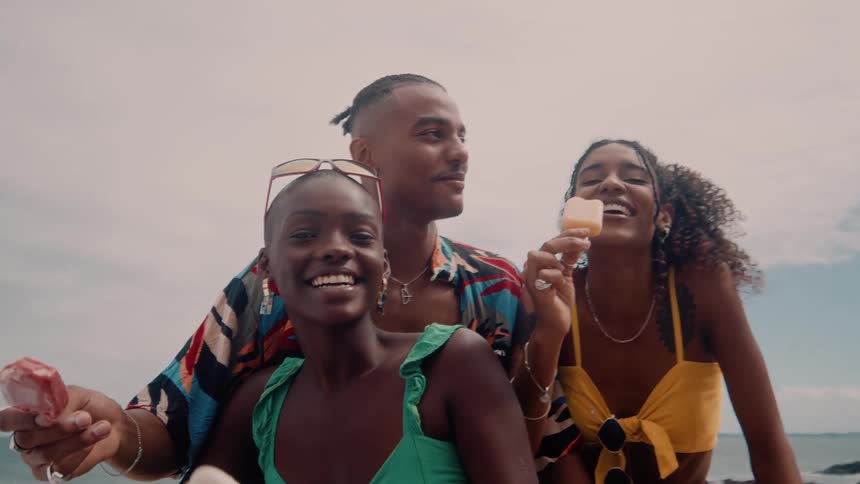 A group of three friends standing on a beach and holding popsicles they are laughing out loud
