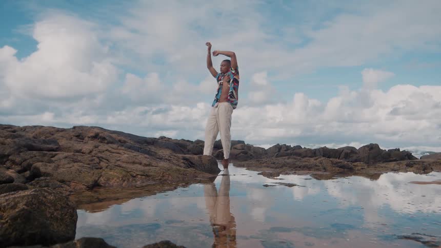 A man wearing a flowery tshirt is standing on the rocks of a beach with a puddle of water in front of him and stretching his arms up