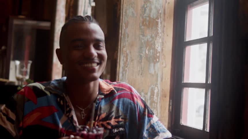 A smiling man is sitting by a window enjoying a drink at a rustic cafe or bar