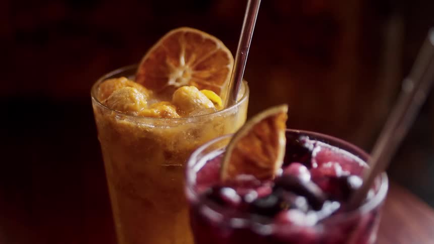 A closeup shot shows two colorful fruit cocktails in tall glasses garnished with dehydrated orange slices in a dimly lit bar