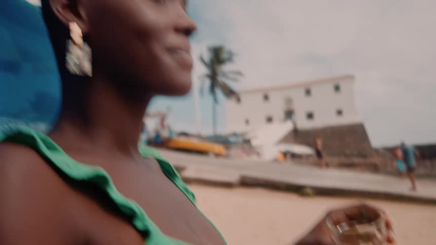 The face of a smiling woman who is sitting in a chair on the beach and holding a glass of beer in her hand