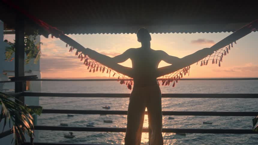 Man holding onto a hammock stands silhouetted against the setting sun on a roof covered balcony