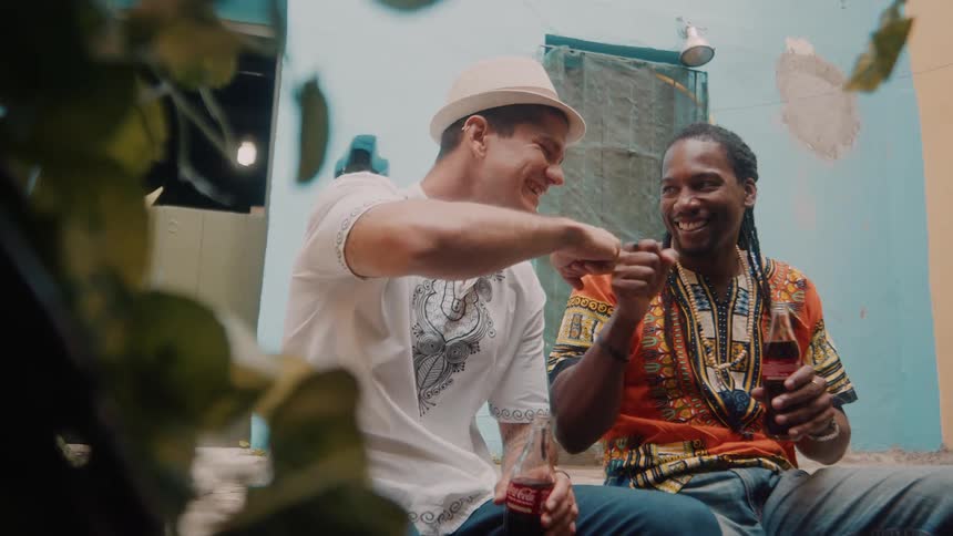Two men laugh and fistbump while enjoying bottled drinks in a colorful outdoor setting