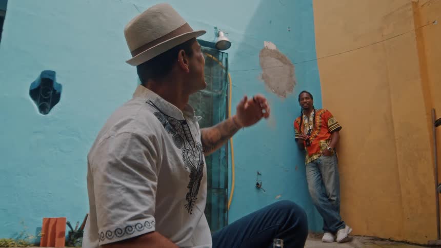 Two men are in a colorful outdoor area one sitting and wearing a hat the other standing and leaning casually against a wall