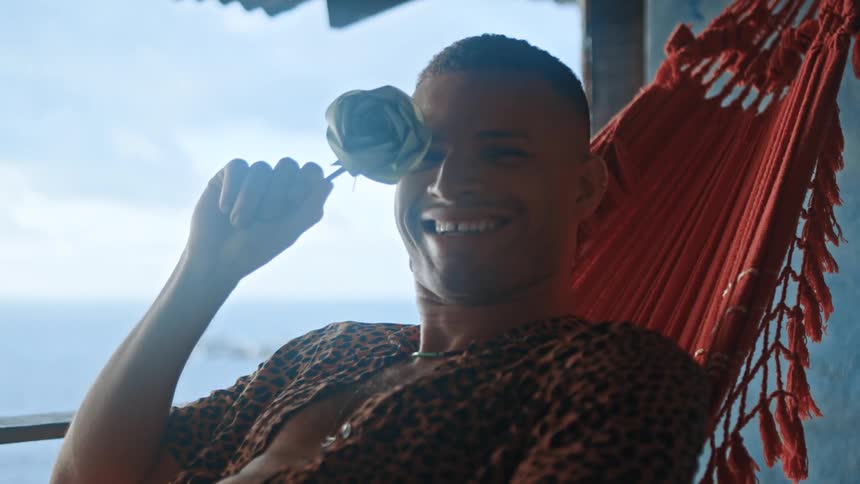 Man on a red hammock laughing while he covers his right eye with a white rose