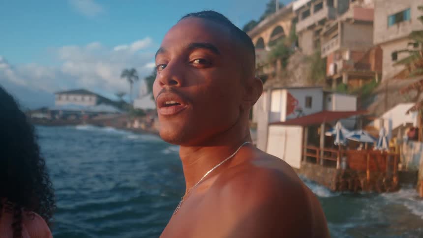 A shirtless man stand near the edge of a rocky coastline with waves crashing below and a coastal town
