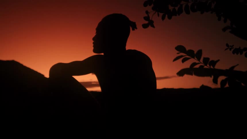 A person sits silhouetted against a vivid orange sunset sky framed by dark tree branches in the foreground