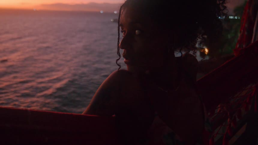 Woman leans against a hammock on a balcony near the coast at sunset