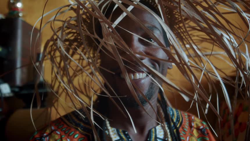 Man smiles and laughs while wearing a hat containing long unbound grass blades