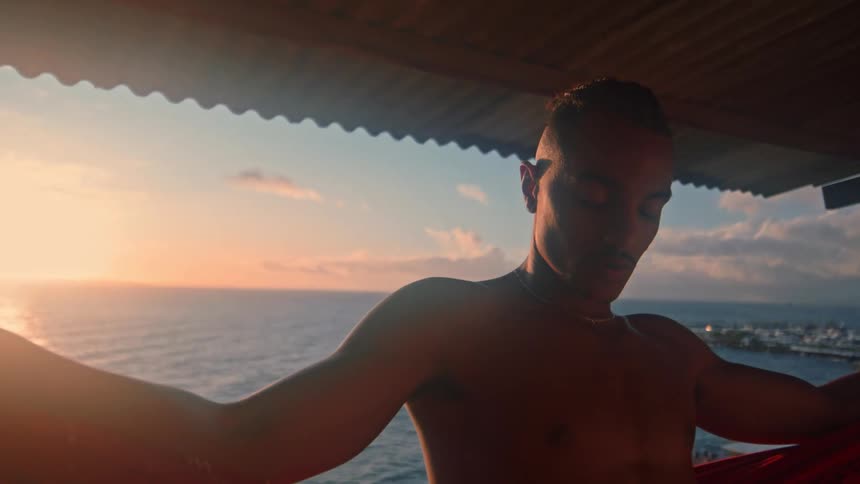 Man stands on a covered balcony with his back to the rising sun and his eyes closed