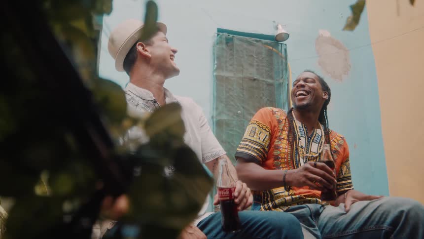 Two men laugh and hold drinks while sitting on a pavement outdoors during daytime