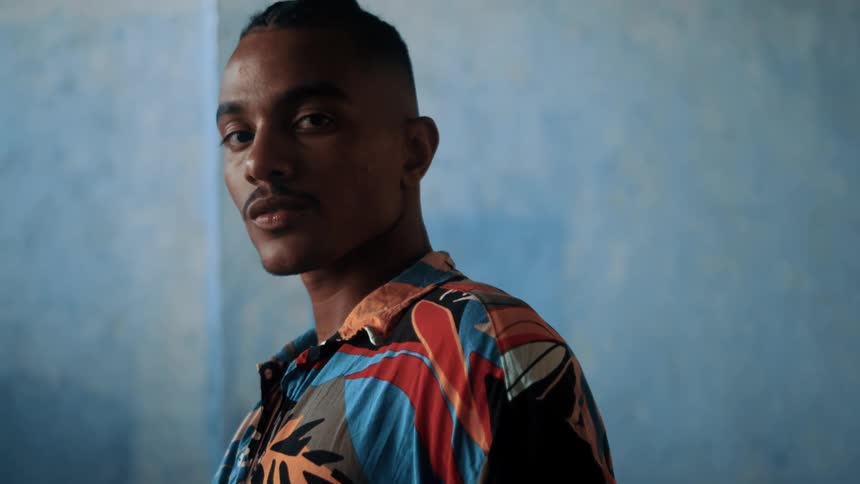 The face of a man wearing a colorful shirt and he is standing looking forward and behind him there is a blue wall