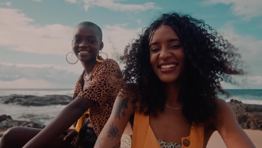 Two joyful women sit on a beach smiling brightly at the camera