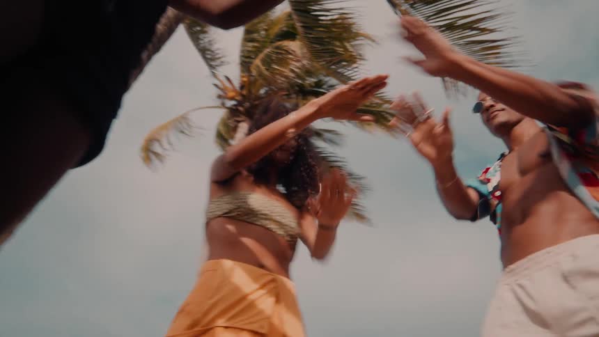 Three people in colorful summer clothes are dancing using their arms in an outdoor setting next to palm trees