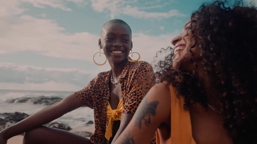Two women sit on the sand near the ocean one in a leopard print shirt and hoop earrings the other in a sleeveless yellow top with curly hair