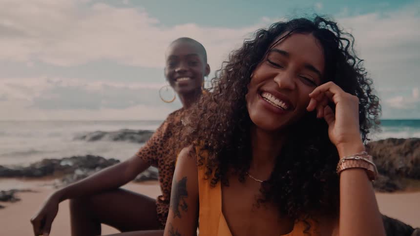 Two women smile at the camera while sitting on a beach near the water