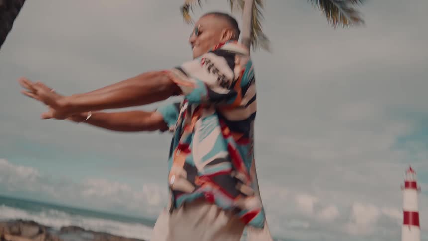 A man and woman dance playfully on a sandy beach with palm trees the ocean and a lighthouse behind them