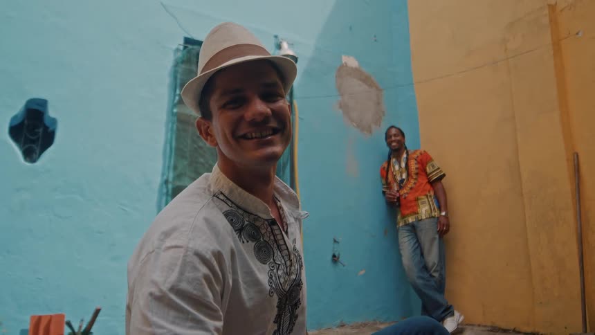 A man in a fedora smiles in a colorful alley while another man leans against a blue wall behind him