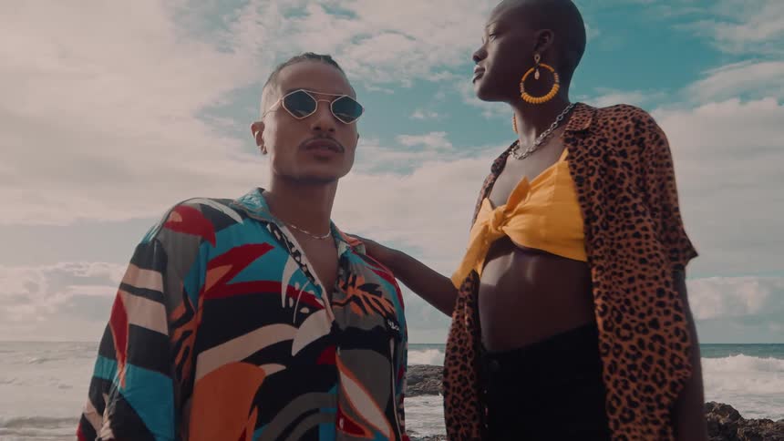 A man and a woman are standing in an environment with a beach in the background and the woman is holding on to the shoulder of the man who is wearing sunglasses