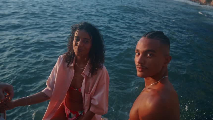A shirtless man and a woman in a pink shirt stand near the edge of a rocky coastline with waves crashing below and a coastal town faintly visible in the distance