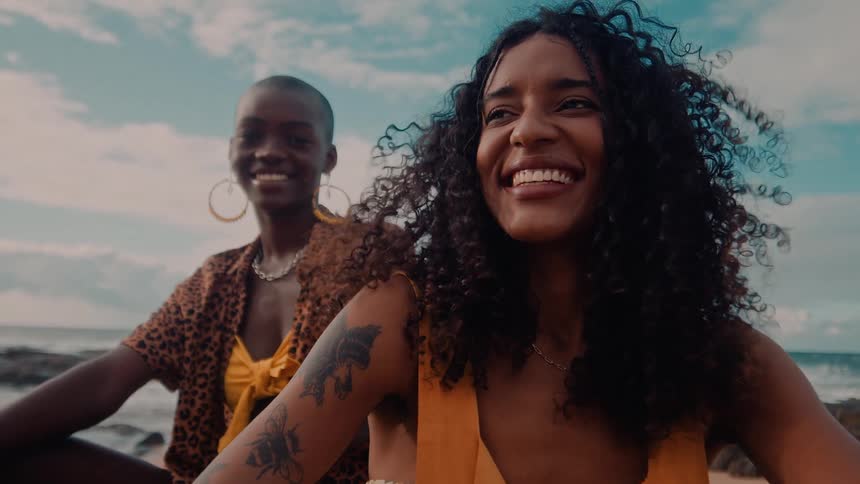 Two women smile and relax on a beach with ocean waves in the background
