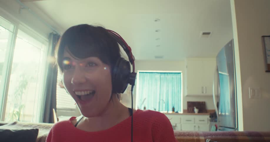 A woman happily plays a video game at home while wearing headphones