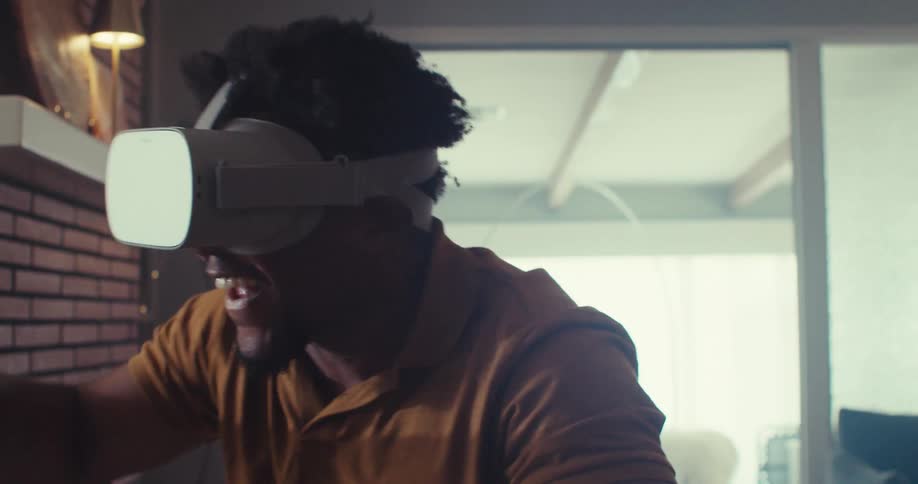 A person wearing a virtual reality headset stands in a room gesturing with both hands with a floor lamp on the left and a couch in the background