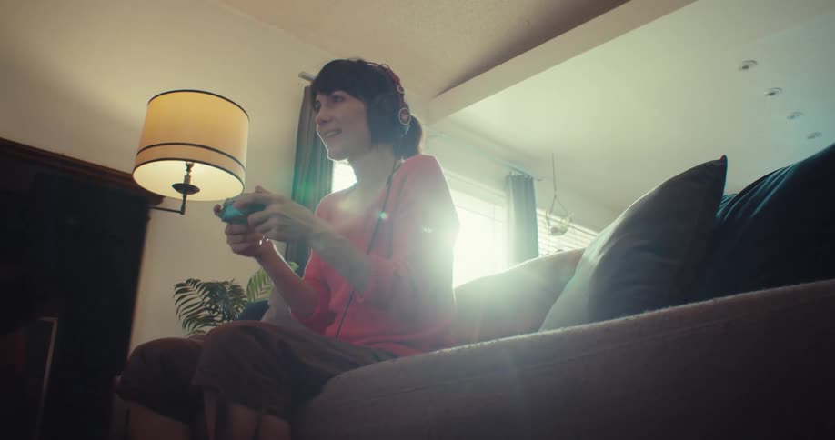 A woman sits on a couch smiling and playing a video game with headphones on