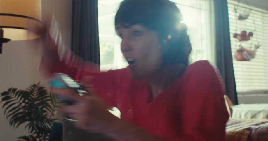 An excited woman wearing headphones laughs and triumphantly pumps her fist while playing a video game in a sunlit room