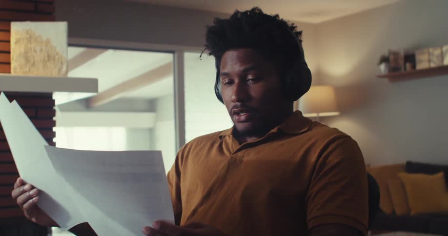A man sits at his desk wearing headphones and shuffling papers in his hands before reading them to himself out loud in his living room on a bright day