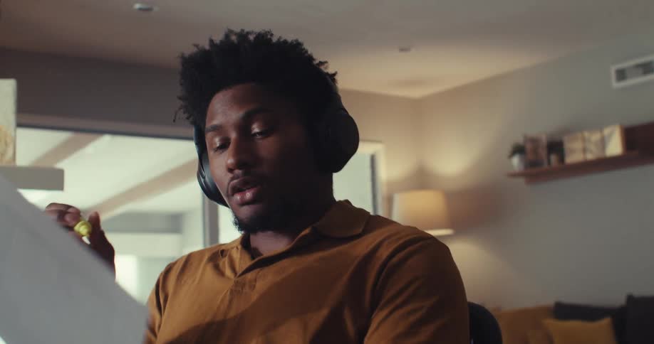 A man in a yellow shirt and headphones reviews a document while sitting in a living room office