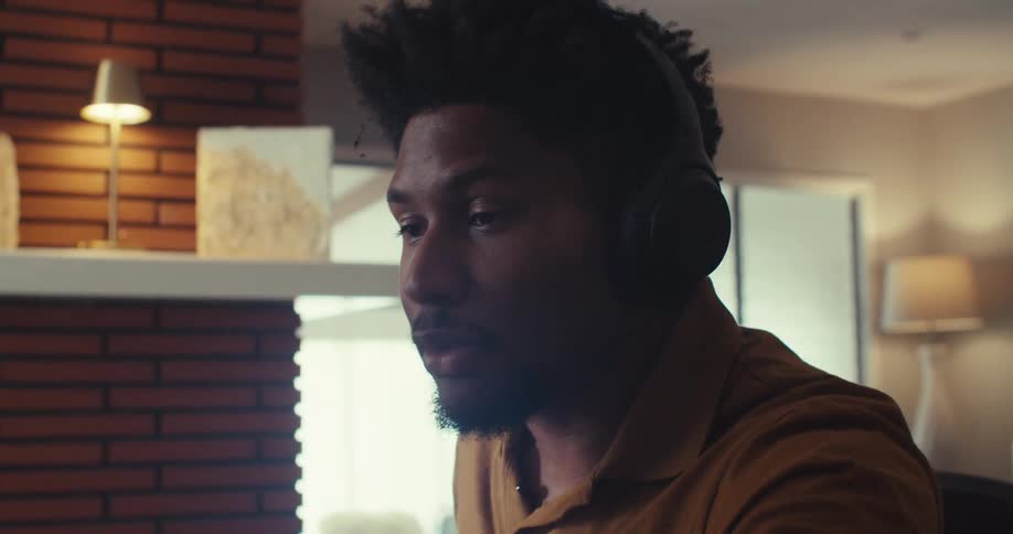A man in a yellow shirt and headphones sits in a modern room speaking to someone off camera or on a video call