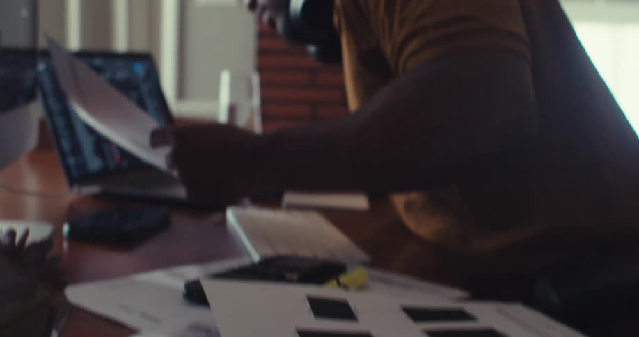 A man examines printed graphs and charts at a desk while referencing stock data on his laptop