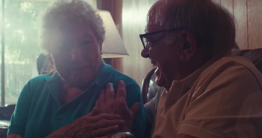 An elderly couple sits on a sunlit couch at home smiling laughing and hugging warmly