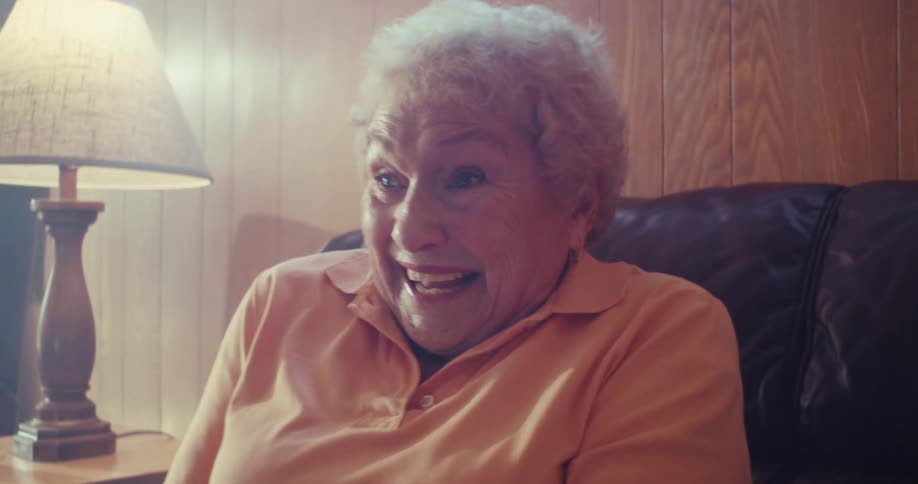 An elderly woman in an orange polo sits on a couch at home enthusiastically playing video games