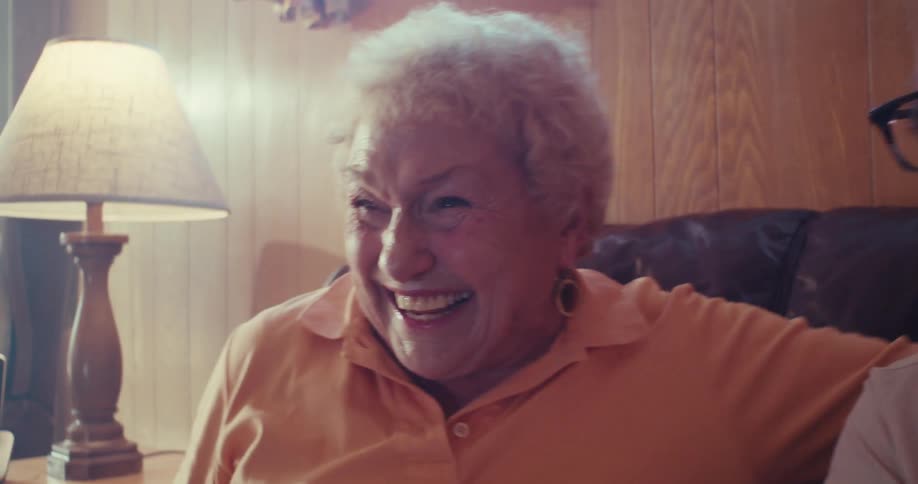 An elderly couple laughs together while sitting on a cozy couch in a warmly lit room