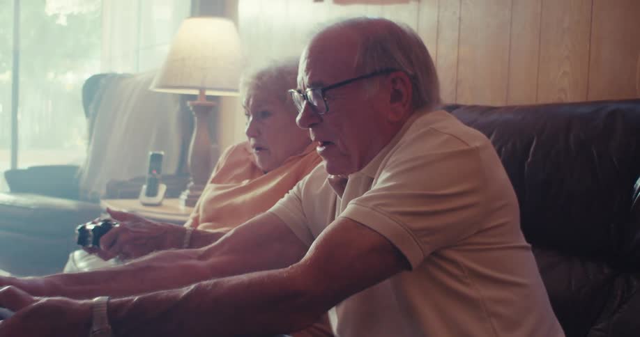 An elderly couple sits on a couch intensely playing video games together in a cozy living room