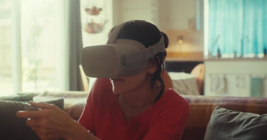 Woman plays a virtual reality game with vr goggles while seating on a couch in a living room during daytime